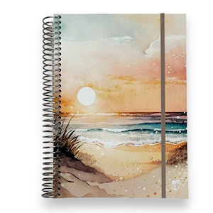 May include: Spiral-bound notebook featuring a watercolor painting of a beach scene at sunset. The artwork depicts a sun, ocean waves, and sand dunes in shades of orange, blue, and beige. A brown elastic band secures the notebook.