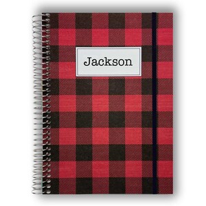 May include: A spiral-bound notebook featuring a red and black buffalo plaid pattern. A white rectangular label is centered on the front cover with the name Jackson printed in black text. A thin black elastic band runs vertically along the right side.