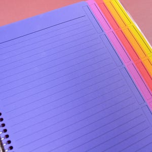 May include: A spiral-bound notebook featuring a purple cover with horizontal lines and a top header section. The right edge displays a multi-colored tabbed index with vertical sections in pink, orange, and yellow, bound by a silver metal coil.