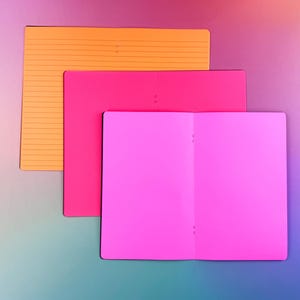 May include: Three notebooks stacked on top of each other. The top notebook is orange with lined paper. The middle notebook is pink. The bottom notebook is a larger pink notebook with a blank page.
