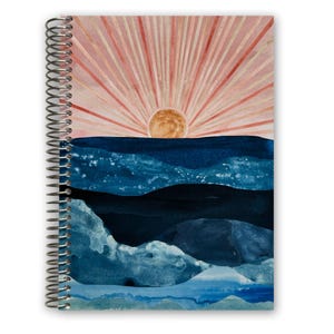 May include: Spiral-bound notebook featuring an abstract watercolor landscape. The design includes a rising sun with radiating lines above blue and navy mountain ranges. The notebook's cover has a watercolor effect with shades of pink, orange, and blue.