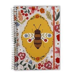 May include: Spiral-bound notebook with a bee illustration. The cover features a large bee in a yellow oval, surrounded by floral patterns in red, pink, and blue on a cream background. A red elastic band secures the notebook.
