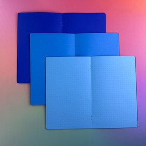 May include: Three open notebooks stacked vertically, each featuring a different shade of blue. The top notebook is dark blue, the middle is medium blue, and the bottom is light blue. Each notebook page displays a consistent dot grid pattern.