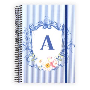 May include: Spiral-bound notebook with a blue and white striped cover. The cover features a blue shield design with the letter "A" in the center, surrounded by floral accents. A blue elastic band secures the notebook.