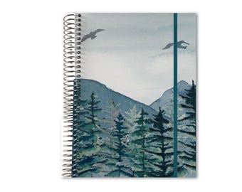 May include: A spiral-bound notebook featuring a watercolor-style landscape illustration of dark green pine trees against blue mountains under a pale sky. Two dark birds are depicted in flight near the top, and a vertical teal elastic band is attached.
