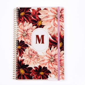 May include: A spiral-bound notebook featuring a floral pattern of deep red and light pink dahlia flowers. A central white oval contains the dark red capital letter M. A light pink elastic band secures the cover vertically on the right side.