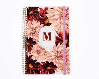 May include: A spiral-bound notebook featuring a floral pattern of deep red and light pink dahlia flowers. A central white oval contains the dark red capital letter M. A light pink elastic band secures the cover vertically on the right side.