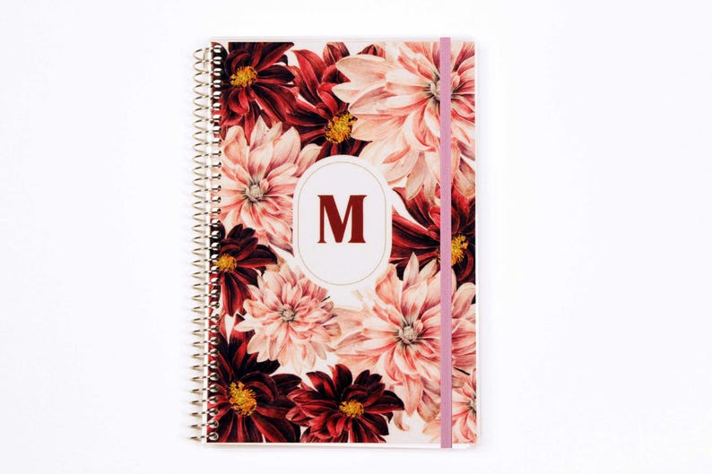 May include: A spiral-bound notebook featuring a floral pattern of deep red and light pink dahlia flowers. A central white oval contains the dark red capital letter M. A light pink elastic band secures the cover vertically on the right side.