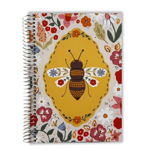 May include: A spiral-bound notebook with a floral and bee design. The cover features a large bee illustration in a yellow oval, surrounded by flowers in red, white, and orange on a cream background.