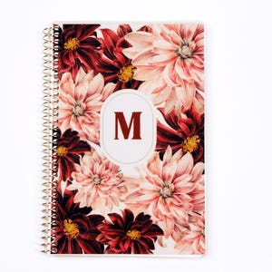 May include: A spiral-bound notebook featuring a floral pattern of deep red, burgundy, and soft pink dahlia flowers. In the center, a white oval frame contains the capital letter M in a dark red serif font, set against a white background.