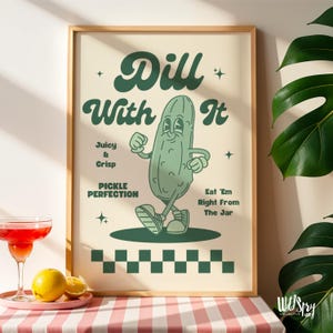 Op de afbeelding: Een ingelijste poster met een cartoon augurkenkarakter en de tekst "Dill With It". De poster heeft een lichtbeige achtergrond met groene tekst en een groene augurkenillustratie. Extra tekst is onder meer "Juicy & Crisp" en "Eat 'Em Right From The Jar."