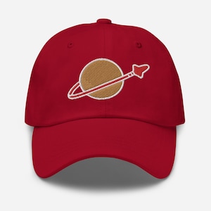 Op de afbeelding: Rode baseballpet met een geborduurd bruin planeet- en rood raketontwerp.