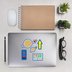 Può includere: Un computer portatile argentato con cinque adesivi colorati sul coperchio. Gli adesivi sono di un pianeta con anelli, un razzo, una freccia che punta verso l'alto, un piccolo pianeta con anelli e un pannello di controllo blu e verde.
