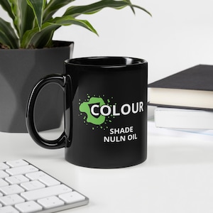 May include: A black ceramic mug with a large handle. The mug features the word "COLOUR" in white, with a green splatter design, and the words "SHADE NULN OIL" in white. The mug is on a white surface.