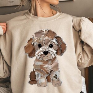 Puede incluir: Sudadera beige con un gráfico de un perro marrón y blanco. La ilustración del perro tiene un diseño de patchwork, con una apariencia texturizada. El perro tiene ojos negros grandes y una nariz pequeña negra. La sudadera es de color neutro.