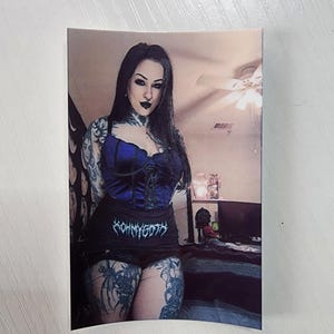 Könnte beinhalten: Foto einer Person, die ein blaues Korsett-Top und schwarze Shorts mit dem Text "KOHMYGOTH" trägt. Die Person hat Tätowierungen an Armen und Beinen. Der Hintergrund zeigt ein Schlafzimmer.