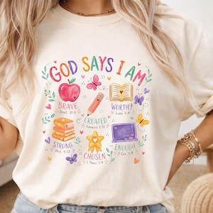 May include: A cream-colored t-shirt featuring a colorful graphic with the text "GOD SAYS I AM" surrounded by illustrations of an apple, a pencil, an open book, a stack of books, a star, and a laptop, along with butterflies and small hearts.