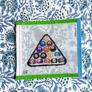 May include: A needlepoint canvas featuring a triangle of billiard balls with numbers and colors. The canvas has a green border and a blue edge. The background is a white mesh. The canvas is on a blue and white floral patterned fabric.
