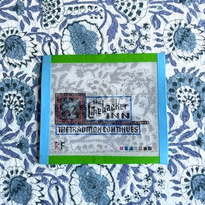 May include: A needlepoint canvas with a blue and green border. The canvas features text that reads "The Cracker Inn" and "The Tradition Continues". The background is a floral pattern in blue and white.