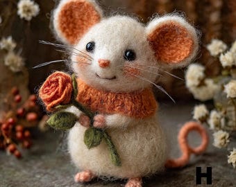 Puede incluir: Una figura de ratón de fieltro hecha a mano con cuerpo crema, orejas y bufanda naranja, y una rosa. El ratón sostiene una rosa con hojas verdes y tiene ojos negros y bigotes. Un objeto decorativo encantador.