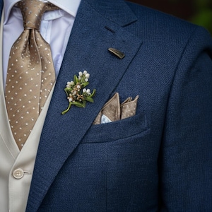 Puede incluir: Primer plano de una chaqueta de traje azul marino con chaleco beige, corbata estampada y prendedor floral. Un pañuelo de bolsillo es visible en el bolsillo del pecho, añadiendo un toque de elegancia al atuendo formal.