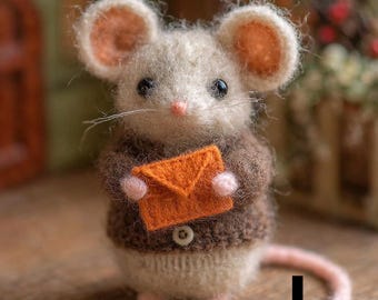 Puede incluir: Una figura de ratón artesanal con cuerpo crema, grandes orejas rosas y un suéter marrón. El ratón sostiene un sobre naranja y tiene una larga cola rosa. Un encantador ejemplo de artesanía hecha a mano.