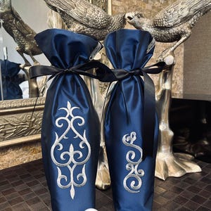 Könnte beinhalten: Zwei Weinflaschentaschen aus dunkelblauem Satin mit silberner Stickerei. Die Taschen sind mit schwarzem Band gebunden und haben den Schriftzug "icdenico" auf der Unterseite.