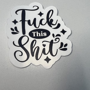 May include: A white sticker with black text that reads "Fuck This Shit." The text is in a stylized font with decorative elements. The sticker has a cloud-like shape.