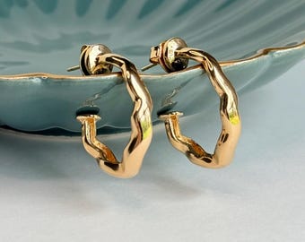 May include: A pair of gold-colored hoop earrings with a textured surface. The earrings are displayed on a teal-colored, scalloped-edge dish with a gold rim. The background is a soft, neutral color.