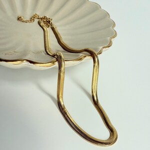 May include: A gold-colored herringbone chain necklace rests on a white, scalloped-edge dish with a gold rim. The necklace is made of small, flat, interlocking links, creating a smooth, flexible design. The background is a solid white.