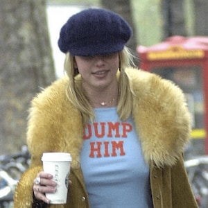 Puede incluir: Una persona con boina azul marino, camiseta celeste con la frase "DUMP HIM" en rojo y abrigo marrón con cuello de piel amarillo. Sostiene una taza de café blanca.