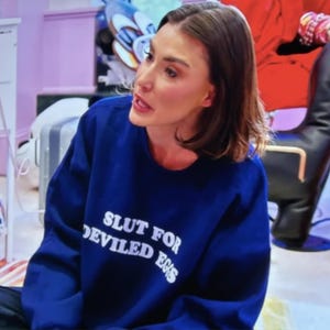 May include: A navy blue sweatshirt with the text "SLUT FOR DEVILED EGGS" in white. The sweatshirt is worn by a person with brown hair. The background includes a black chair and other objects.