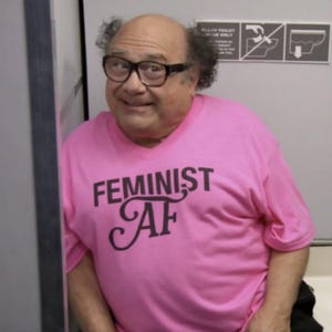May include: A person wearing a pink t-shirt with the words "FEMINIST AF" printed in black. The person is wearing black-framed glasses and is smiling. The background is a light gray wall with a sign.