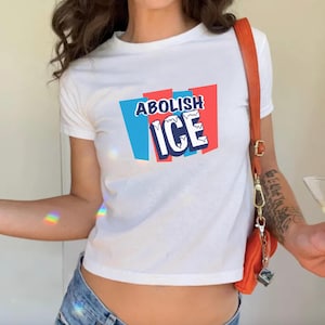 Könnte beinhalten: Weißes Crop-Top-T-Shirt mit dem Text "ABOLISH ICE" in blauen und weißen Buchstaben, vor einem roten und blauen Hintergrund. Das Shirt ist kurzärmlig und hat einen Rundhalsausschnitt.