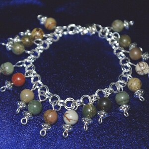 Peut inclure: Un bracelet en cha&icirc;ne argent avec plusieurs breloques pendantes. Les breloques sont faites de diff&eacute;rentes pierres dans des tons de vert, marron et blanc.