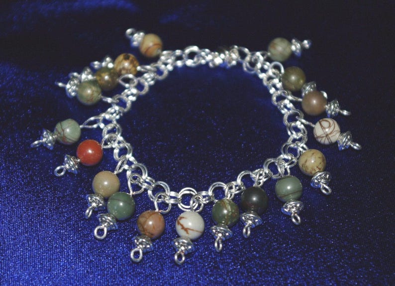Peut inclure: Un bracelet en cha&icirc;ne argent avec plusieurs breloques pendantes. Les breloques sont faites de diff&eacute;rentes pierres dans des tons de vert, marron et blanc.