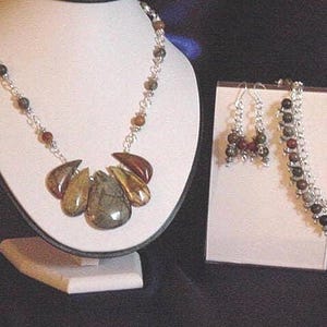 Peut inclure: Un collier en argent avec un pendentif compos&eacute; de plusieurs pierres brunes, vertes et dor&eacute;es. Le collier est pr&eacute;sent&eacute; sur un support blanc. Une paire de boucles d'oreilles et un bracelet avec des pierres similaires sont &eacute;galement pr&eacute;sent&eacute;s.
