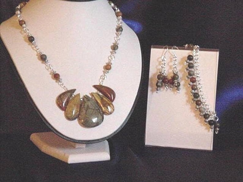 Peut inclure: Un collier en argent avec un pendentif compos&eacute; de plusieurs pierres brunes, vertes et dor&eacute;es. Le collier est pr&eacute;sent&eacute; sur un support blanc. Une paire de boucles d'oreilles et un bracelet avec des pierres similaires sont &eacute;galement pr&eacute;sent&eacute;s.