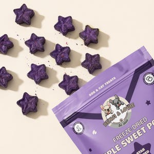 May include: Purple star-shaped dog and cat treats scattered on a white surface. A partially visible bag of Benji & Louie freeze-dried apple sweet potato treats is in the image. The bag has a purple background and the words "Dog & Cat Treats".