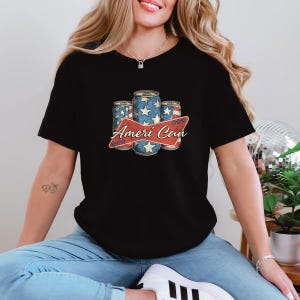 May include: Black t-shirt featuring a graphic of three beverage cans with a patriotic design. The cans have a blue background with white stars and are labeled "Ameri Can" in a red banner.
