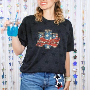 May include: Dark gray t-shirt with a star pattern and a graphic of three beverage cans with a red banner that reads "Ameri Can". The cans have a blue and white star design. The person is wearing blue jeans.