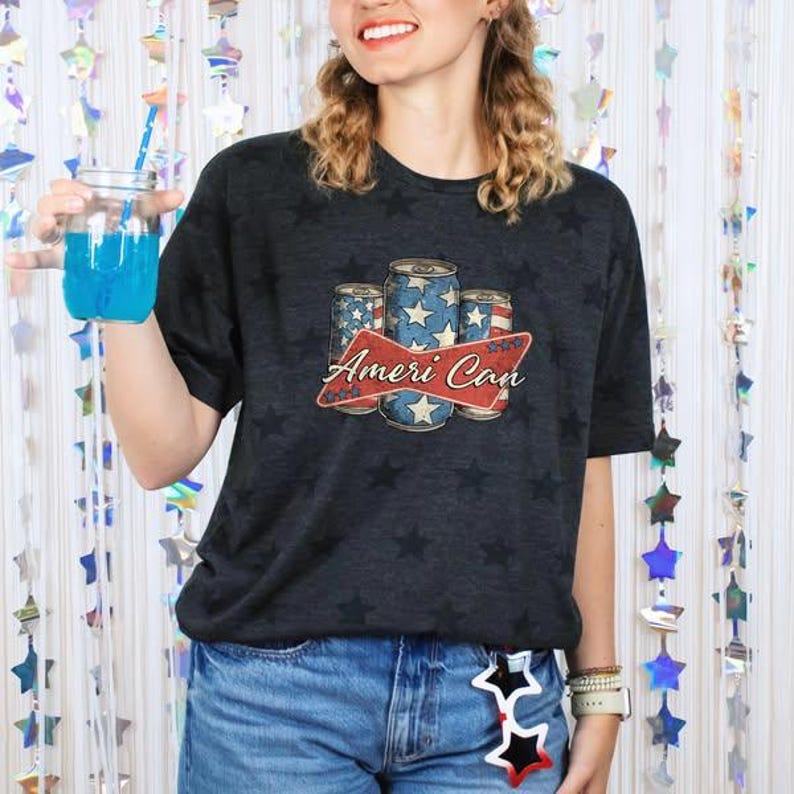 May include: Dark gray t-shirt with a star pattern and a graphic of three beverage cans with a red banner that reads "Ameri Can". The cans have a blue and white star design. The person is wearing blue jeans.