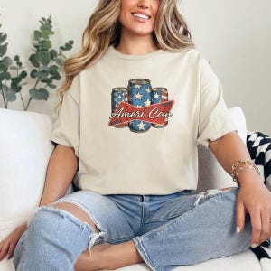 May include: Cream-colored t-shirt featuring a graphic of three beverage cans with a patriotic design of stars and stripes. The text "Ameri Can" is displayed on a red banner. The shirt is worn by a person.