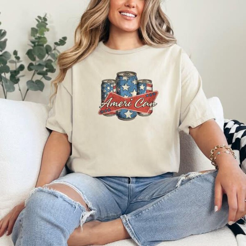 May include: Cream-colored t-shirt featuring a graphic of three beverage cans with a patriotic design of stars and stripes. The text "Ameri Can" is displayed on a red banner. The shirt is worn by a person.