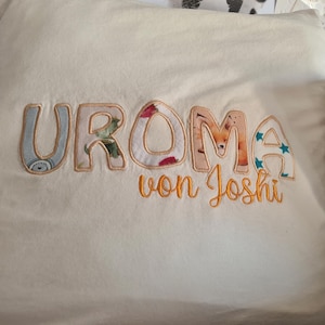 Könnte beinhalten: Ein weißer Stoff mit dem Wort "UROMA" in bunten Stoffbuchstaben mit orangefarbener Naht. Die Worte "von Joshi" sind darunter in Orange gestickt.