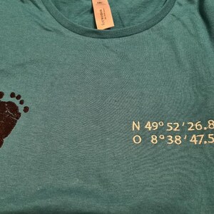 Puede incluir: Camiseta de color turquesa con una huella de pie marr&oacute;n oscuro en el lado izquierdo. Las coordenadas N 49&deg;52'26.868 y O 8&deg;38'47.547 est&aacute;n bordadas en el lado derecho. Cuello redondo.