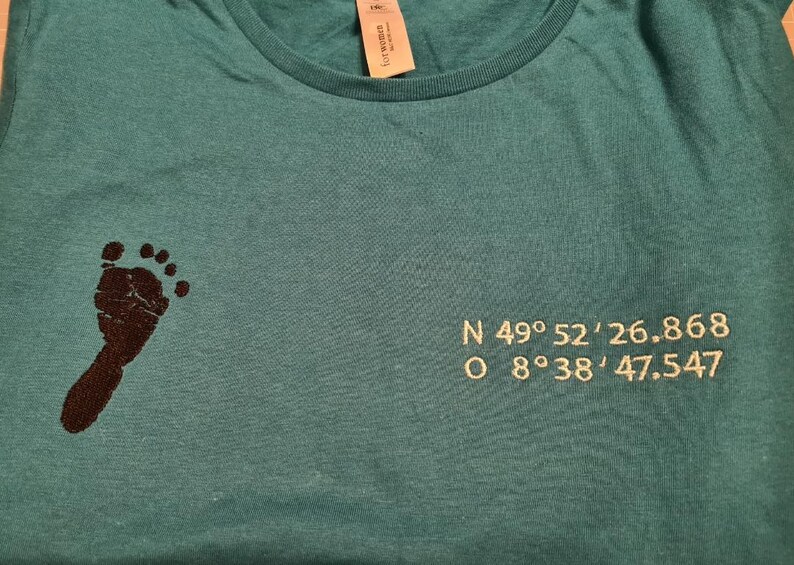 Puede incluir: Camiseta de color turquesa con una huella de pie marr&oacute;n oscuro en el lado izquierdo. Las coordenadas N 49&deg;52'26.868 y O 8&deg;38'47.547 est&aacute;n bordadas en el lado derecho. Cuello redondo.