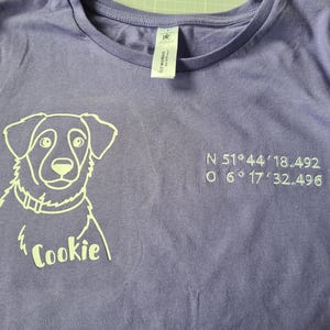Könnte beinhalten: Lilafarbenes T-Shirt mit weißer Umrisszeichnung eines Hundes und dem Namen "Cookie" darunter. Auf der rechten Seite sind Koordinaten gestickt. Das Oberteil hat einen Rundhalsausschnitt und kurze Ärmel.