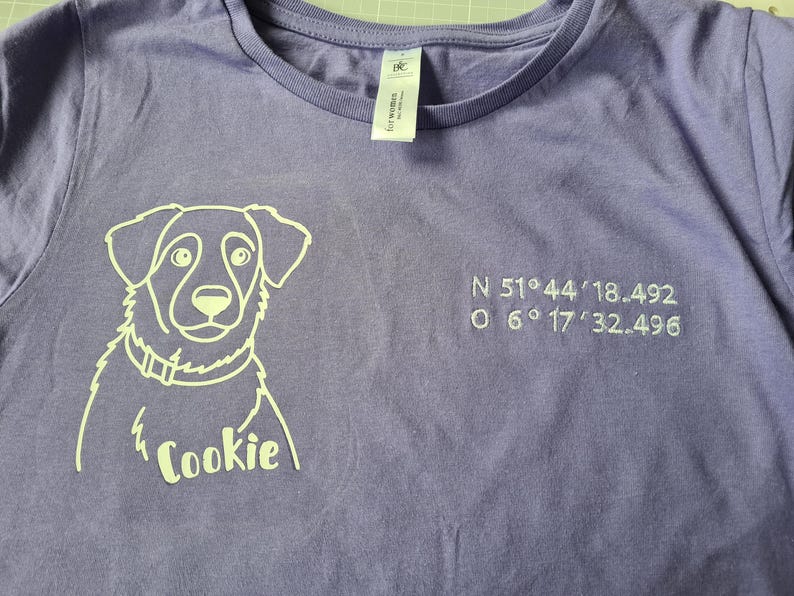 Puede incluir: Camiseta lila con un contorno blanco de un perro y el nombre "Cookie" debajo. Coordenadas bordadas a la derecha. La camiseta tiene cuello redondo y mangas cortas.