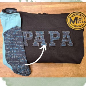 Könnte beinhalten: Schwarzer Kapuzenpullover mit einem blauen und weißen "PAPA"-Design aus Code. Der Code ist in hellblauer Schrift auf dunkelblauem Hintergrund. Der Pullover hat ein kleines Etikett mit der Aufschrift "Handmade Micki Makes with love".